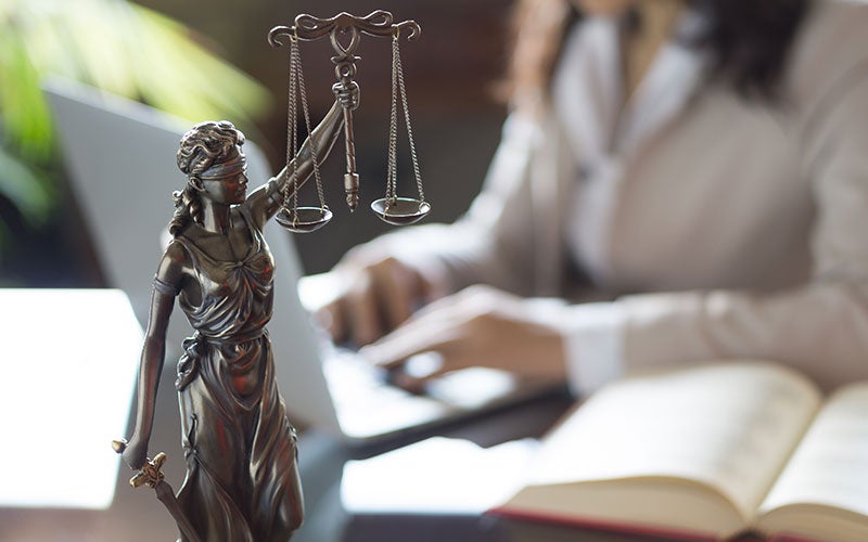 Bronze statue of Lady Justice with scales, in front of a blurry person typing on a laptop and an open book.
