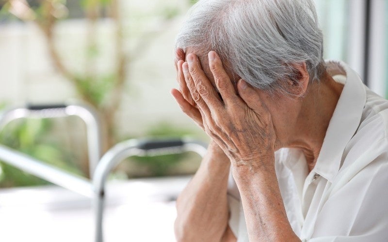 An elderly person with gray hair covers their face with their hands, appearing distressed, with a walker in the blurred background.